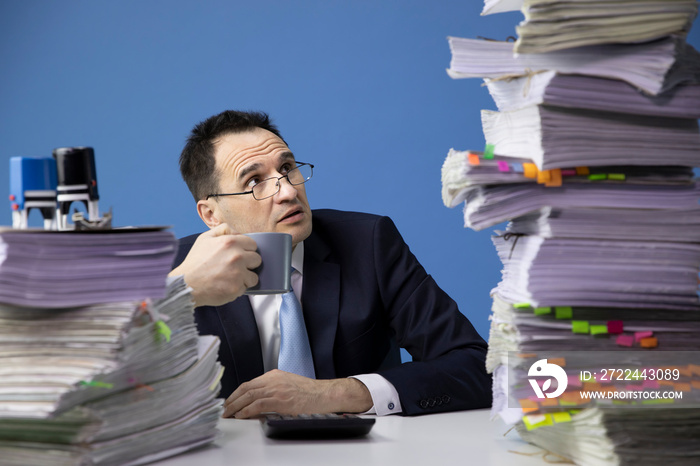 knackered office employee looks frightened at high stack of documents. Business man employee being desperate of too much work at his desk full of papers. overhour job concept. isolated on blue