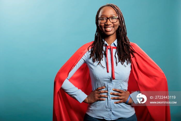 Proud and happy young superhero woman posing brave for camera while standing on blue background. Strong and selfless justice defender wearing mighty hero red cloak while standing with hands on hip.