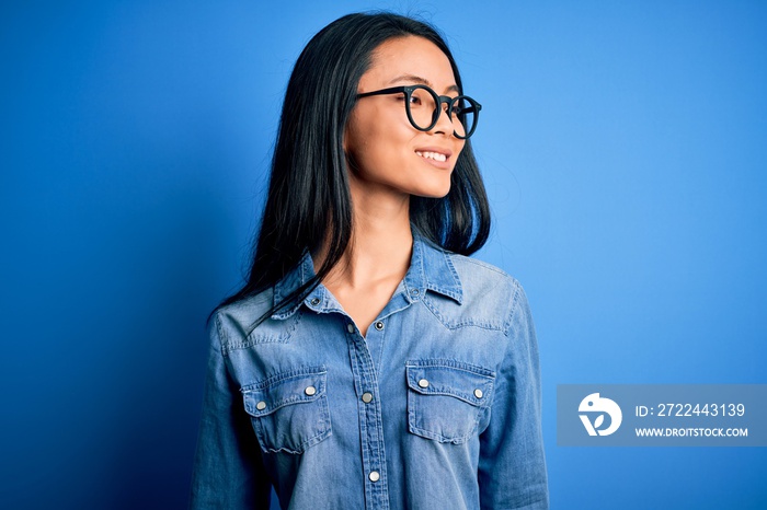 Young beautiful chinese woman wearing casual denim shirt over isolated blue background looking away to side with smile on face, natural expression. Laughing confident.