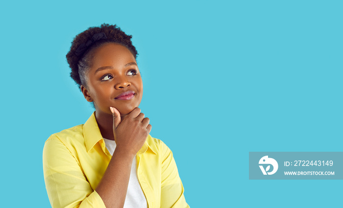 Young woman dreaming or thinking about something. Happy beautiful young Afro American girl in casual yellow shirt standing isolated on blue background, holding hand on chin and looking at copy space