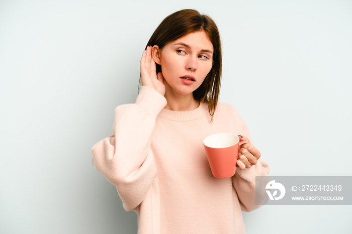 Young English woman holding a mug isolated on blue background trying to listening a gossip.