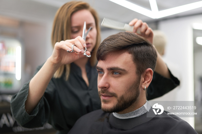 Handsome blue eyed man sitting in barber shop. Hairstylist Hairdresser Woman cutting his hair. Female barber.