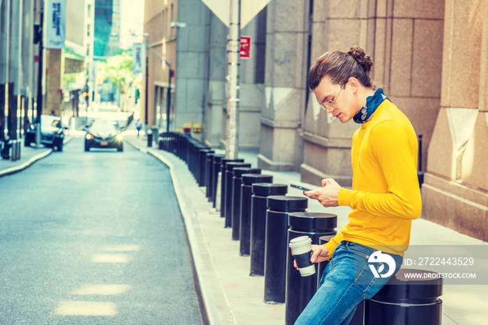 Working Break. Young Hispanic American with hair bun, wearing glasses, yellow long sleeve T shirt, blue jeans, small black scarf around neck, holding cup of coffee, sitting on street, texting..