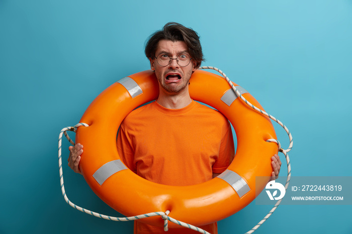 Depressed man lifeguard cries with panicking expression, poses with inflated swimming ring, tired of saving drowning people, isolated on blue background. Hopeless desperate male cannot swim well