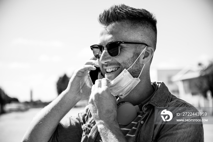 Smiling man with headphones around his neck and lowered surgical  mask talking after coronavirus outbreak - Young man talking with mobile phone outdoors after covid-19 lockdown - New normal concept