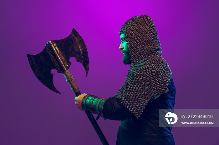 Studio shot of young bearded man in image of medieval warrior, archery in chain armor with ax isolated over purple background in neon. Comparison of eras