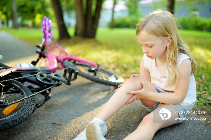 Cute little girl sitting on the ground after falling off her bike at summer park. Child getting hurt while riding a bicycle.