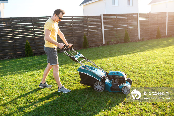 A young man is going to mow the lawn, he starts a push lawn mower. A guy in casual yellow t-shirt and in sunglasses