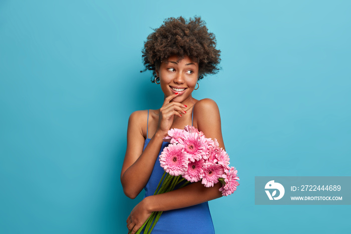 Spring and holiday concept. Smiling glad dark skinned woman has Afro hair holds beautiful bouquet of pink gerberas looks away happily receieves flowers on date with boyfriend celebrates 8 match