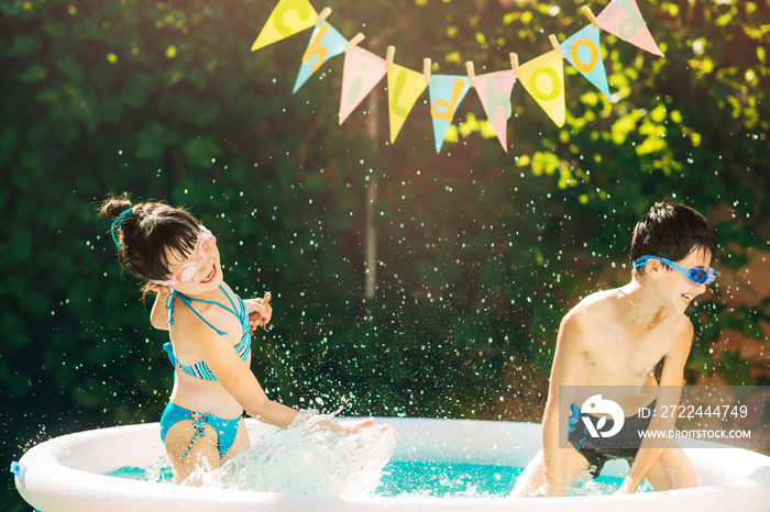 Two young children brother and sister splashing water in inflatable swimming pool summertime sunny day in the yard. Children wearing swimming goggles. Movement, positive emotions, joy.