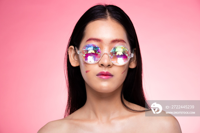 Fashion Model looks at camera for shooting new collection Sunglasses.  Beautiful Asian Woman summer trend make up wear Kaleidoscope Glasses with colorful, studio lighting pink background