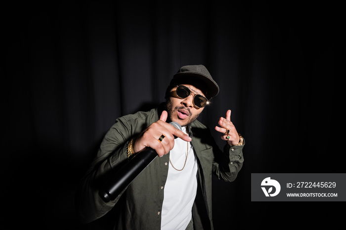 indian hip hop singer in sunglasses and cap singing in microphone on black.