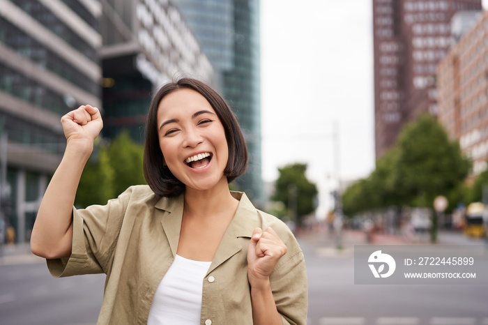 Happy asian girl winning and rejoicing, celebrating on streets of city, dancing from joy, triumphing and smiling