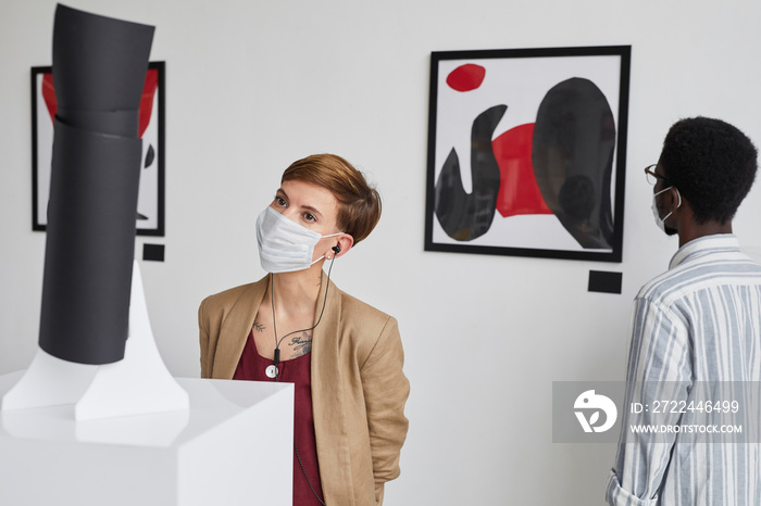 Portrait of two young people looking at paintings and sculptures while wearing masks and exploring modern art gallery exhibition, copy space