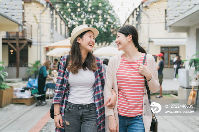 Having fun together on summer holidays. two young cheerful asian korean girls walking enjoying looking at each other and laughing on street little village. sisters relax in creative market thailand