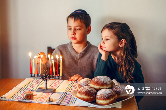 Brother and sister siblings lighting candles on menorah for Jewish Hanukkah holiday at home. Children celebrating Chanukah festival of lights. Dreidel and Sufganiyot donuts in plate on table