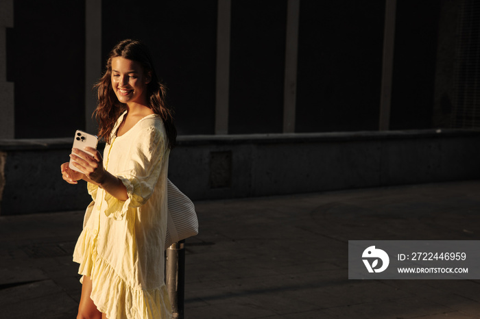 Smiling young caucasian woman is using smartphone relaxing outdoors. Brunette wears casual summer clothes. Online entertainment concept