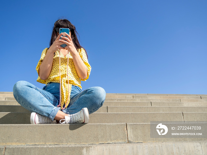 Woman using smart phone outside sitting on stairs addicted to social media. Young brunette girl unrecognized on a summer day typing on phone with pop socket. Low angle with copy space.
