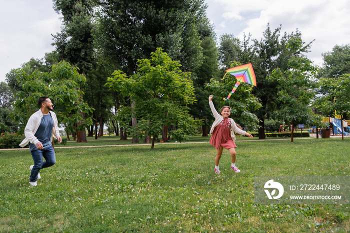 Cheerful asian girl playing with colorful flying kite near dad running in park.