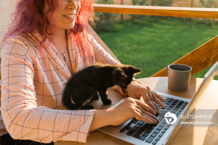 Work from home or study online and videocall with a woman gardener working at laptop outdoor on terrace with kitten close-up, sitting on ratang chair around houseplant and flower pots