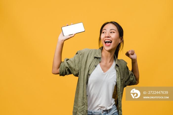 Young Asia lady show empty smartphone screen with positive expression, smiles broadly, dressed in casual clothing feeling happiness on yellow background. Mobile phone with white screen in female hand.