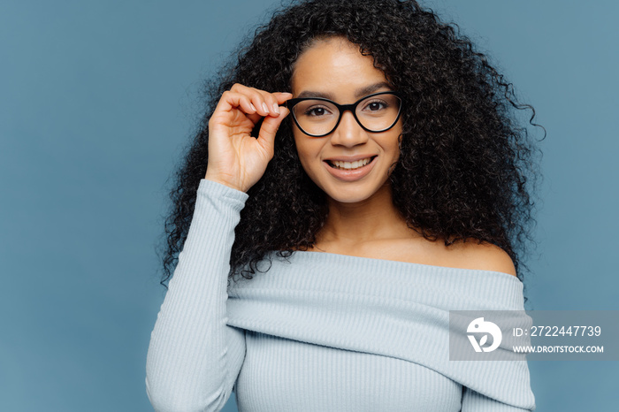 Headshot of pleasant looking young African American woman keeps hand on frame of glasses, has gentle smile, wears blue jumper, has minimal makeup, healthy skin, isolated over blue background