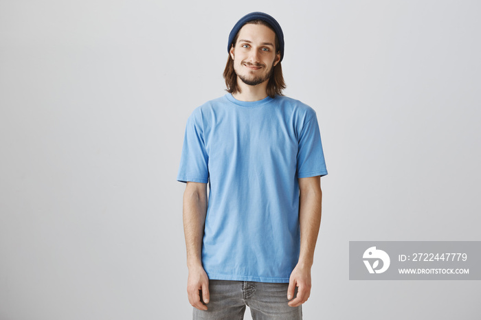 Handsome urban guy likes skating and hanging out. Indoor shot of attractive hipster man in blue beanie and t-shirt, smiling shy and standing casually over gray background, expressing friendliness