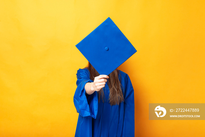 Young woman covering her face with graduating cap over yellow background