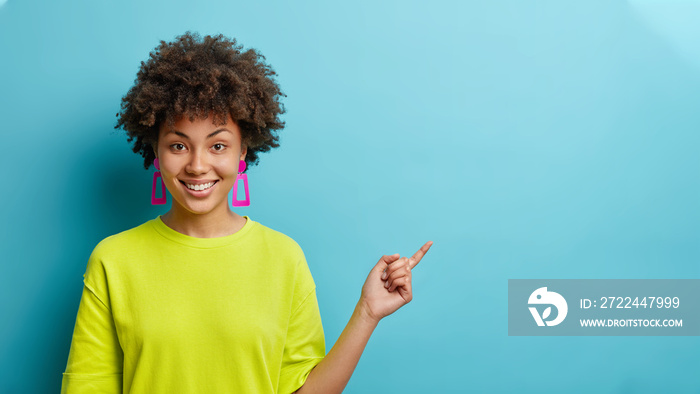 Horizontal shot of cheerful Afro American woman in casual t shirt points away on copy space suggests follow this direction or click on link poses against blue background. Your advertising here