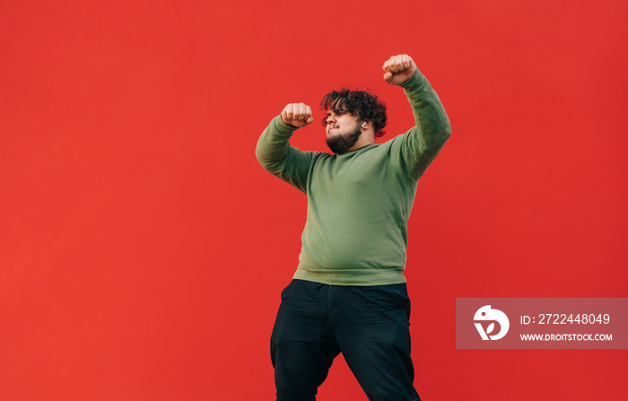 Curly overweight young man in casual clothes dancing and listening to music on headphones, isolated. Fat man shows a dance performance on the street on a background of a red wall.