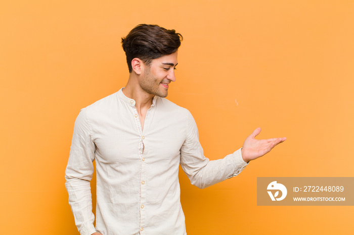 young handsome man feeling happy and cheerful, smiling and welcoming you, inviting you in with a friendly gesture against orange background