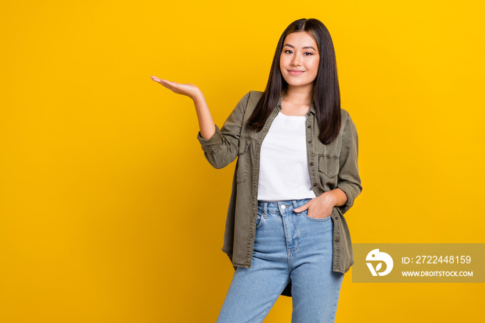 Portrait of cheerful optimistic pleasant girl dressed khaki shirt arm in pocket presenting empty space isolated on yellow color background