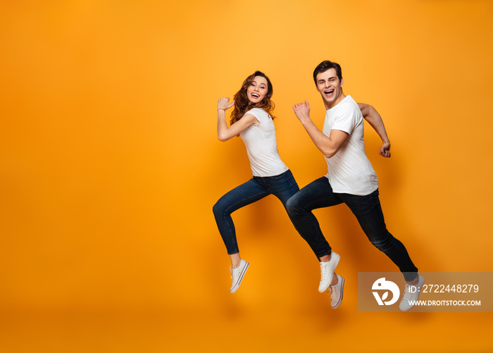 Young lovely cheerful couple posing together on camera while running or jumping, along yellow background
