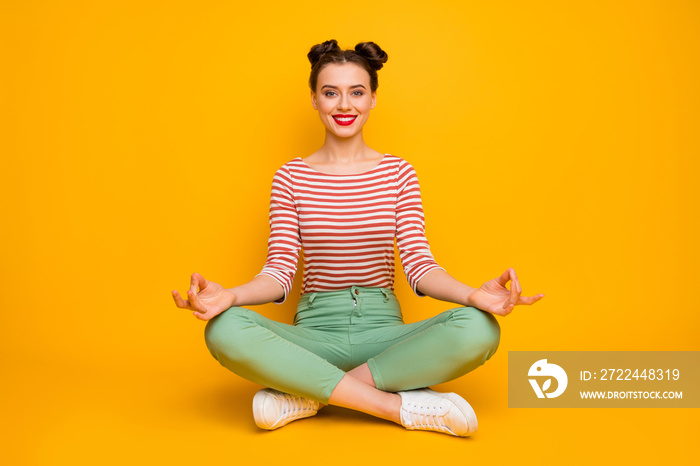 Full length photo of funny lady hold fingers together sit floor legs crossed lotus position wear striped red white shirt green pants footwear isolated yellow color background