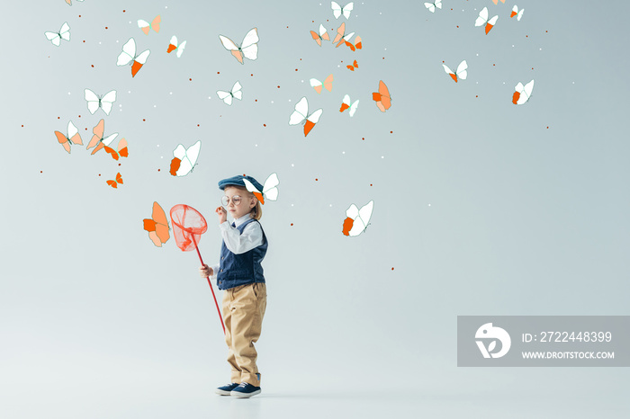 cute and blonde kid in retro vest and cap holding butterfly net on grey background with fairy butterflies