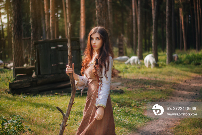 Young beautiful girl in medieval cowboy clothes, with a stick in hand. Barefoot on the ground. Against the background of the forest and green grass. A model with clean skin.