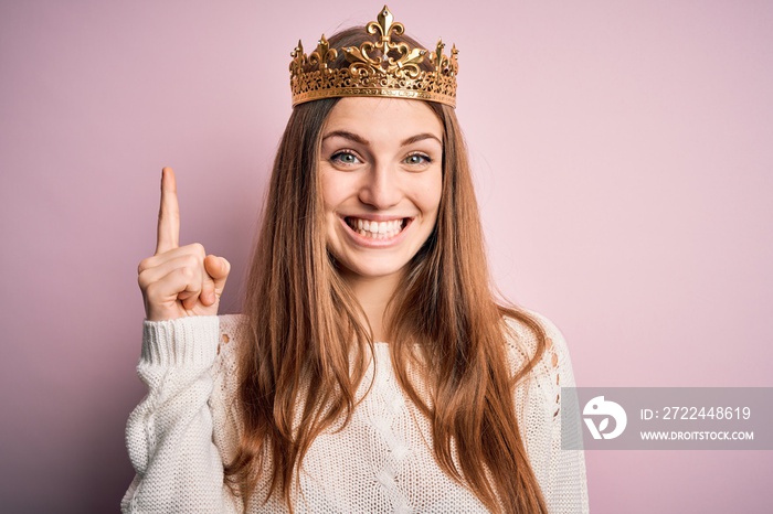 Young beautiful redhead woman wearing queen crown over isolated pink background pointing finger up with successful idea. Exited and happy. Number one.