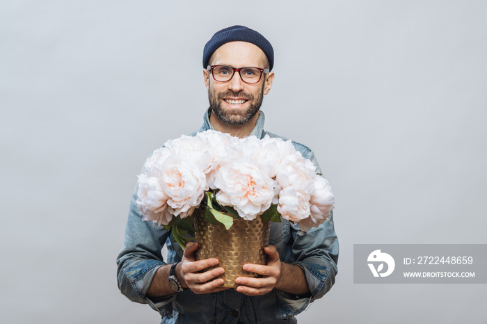 Positive unshaven male wears spectacles, holds beautiful bouquet of flowers, going to present them to his wife and congratulate with birthday, isolated over white background. People, presents concept