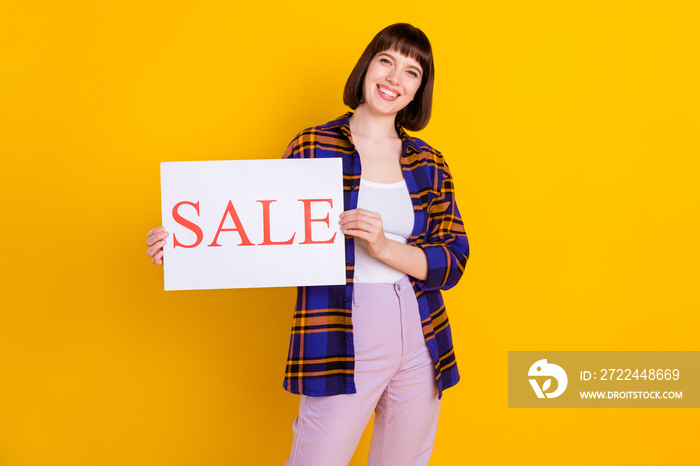 Portrait of attractive cheerful girl wearing plaid shirt holding in hands sale word placard isolated over bright yellow color background