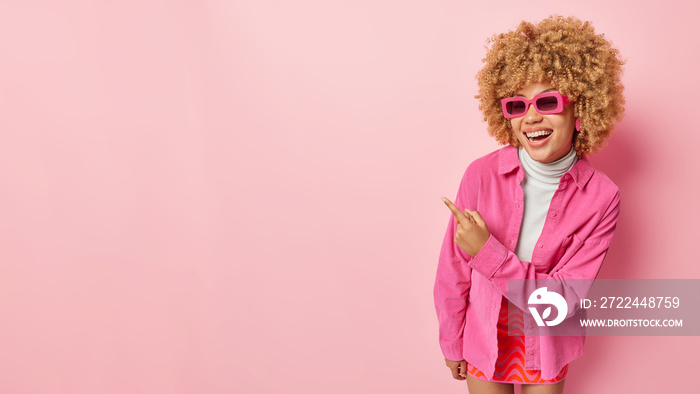 Overjoyed stylish woman wears sunglasses shirt and skirt points finger on right at blank space demonstrates something funny feels very glad isolated over pink background blank space for your advert