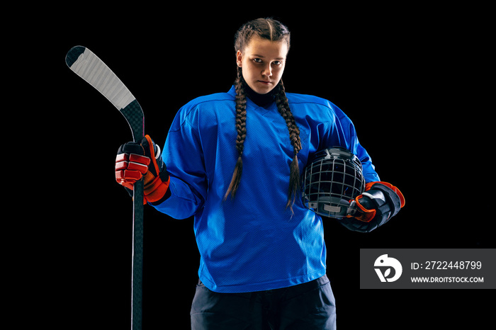 Young female hockey player isolated on black background. Sportswoman wearing equipment and helmet standing with the stick. Concept of sport, healthy lifestyle, motion, movement, action.