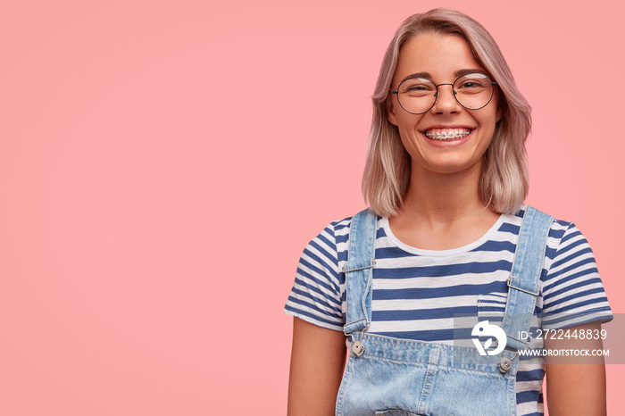 Charming pleasant looking female in stylish denim dungaree and spectacles, listens carefully to somebody`s point of view, expresses positiveness, stands against pink background with copy space