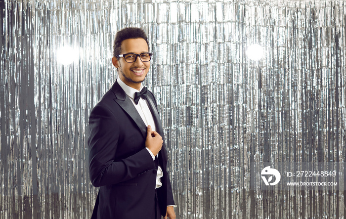 Portrait of attractive and elegant showman or host of festive event on silver shiny background. Smiling young dark-skinned man in black classic suit looks confidently at camera. Banner.