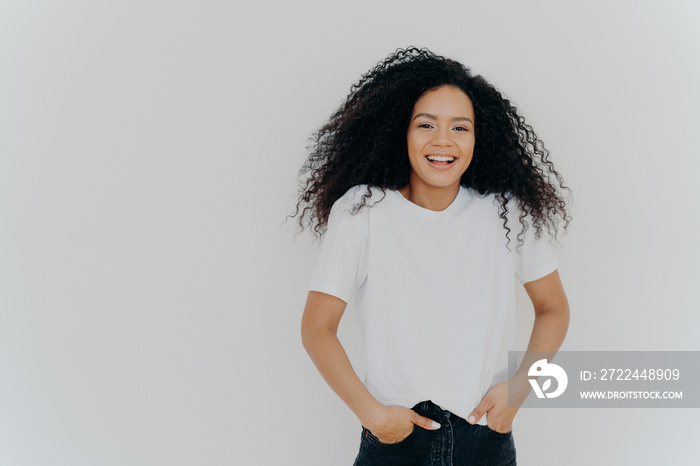 Half length shot of good looking smiling woman laughs at funny joke, has fun, keeps both hands in pockets of jeans, wears white t shirt, has curly fluffy hair, poses indoor, blank space on right side