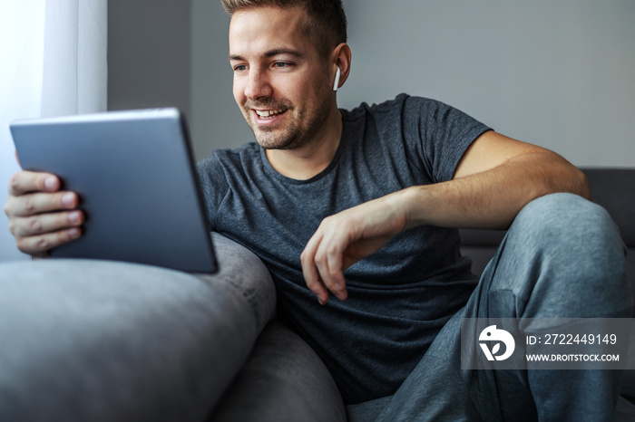 Online chat with friends and family. Isolation, casual weekend. Young and handsome man in sportswear sits comfortably on a gray sofa and holds a tablet in his hands and has white Blue tooth headphones