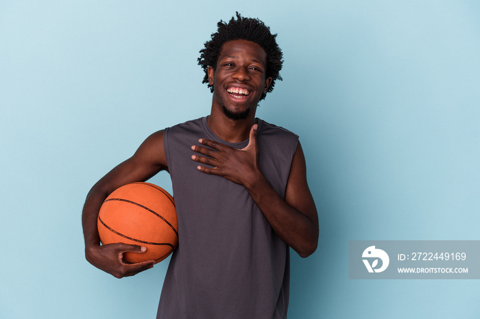 Young african american man playing basketball isolated on blue background laughs out loudly keeping hand on chest.