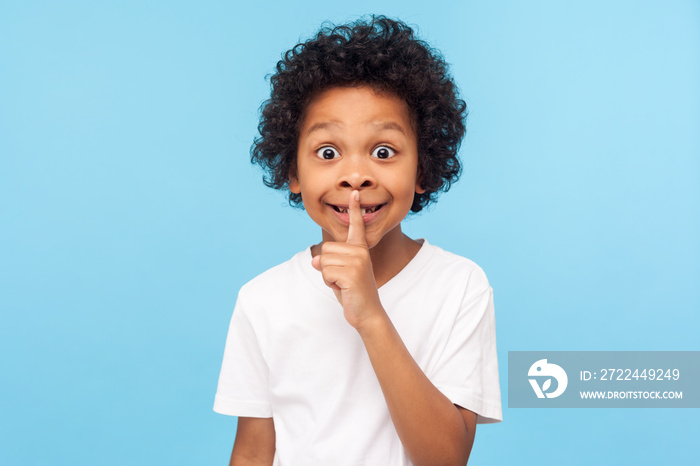 Shh, be quiet! Portrait of funny cute little boy with curly hair in T-shirt making silence gesture with finger on his lips, keeping some secret, child mystery. studio shot isolated on blue background