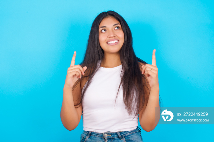 Successful friendly looking Young hispanic girl wearing tank top over blue background exclaiming excitedly, pointing both index fingers up, indicating something.