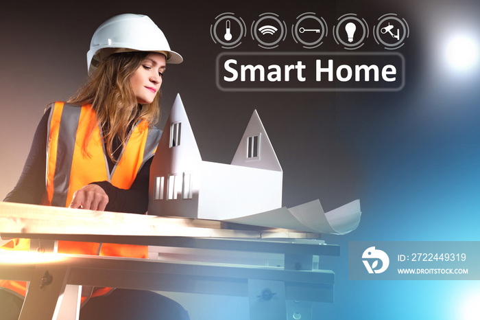 Smart home logo next to  architect. The girl near to layout of cottage. Dark background behind the architect. Concept - she plans to install a smart home system. Woman in orange vest and helmet.