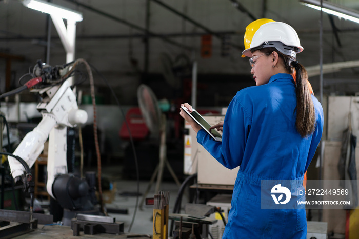 Female automation engineers wear a blue uniform with helmet safety inspection control a robot arm welding machine with a tablet in an industrial factory. Artificial intelligence concept.